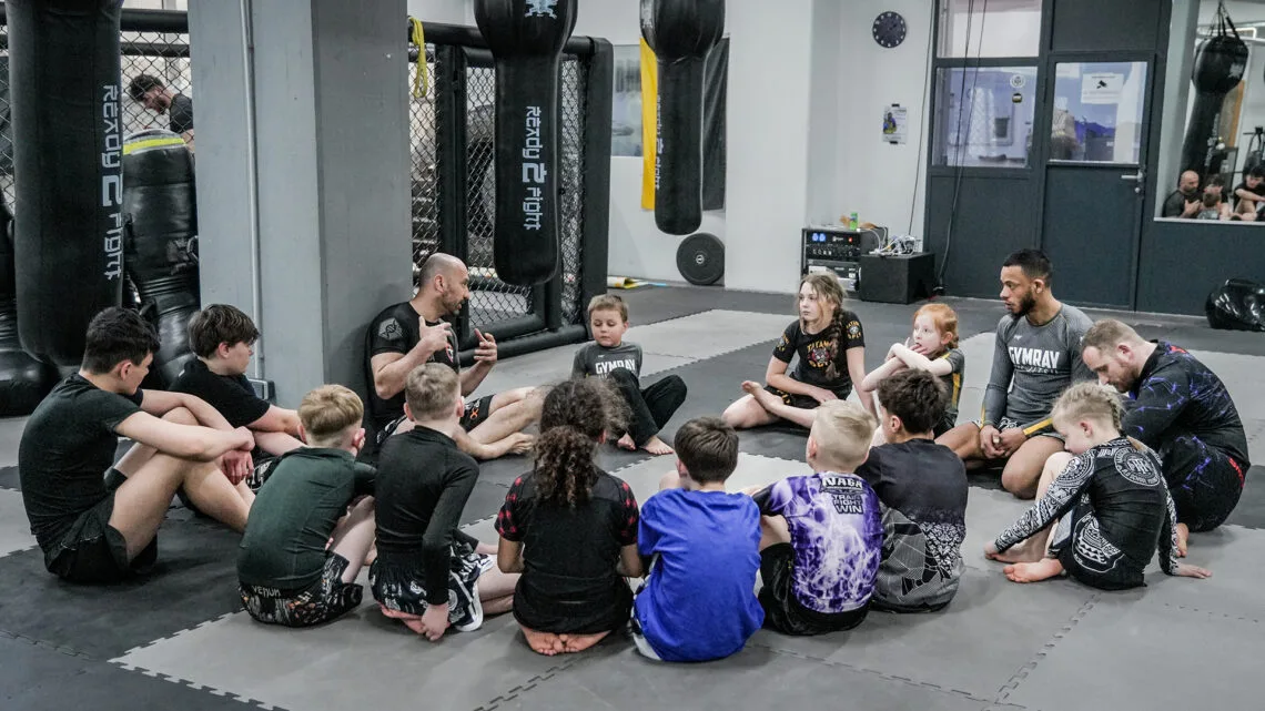 Kinder und Jugendliche sitzen mit ihren Trainern beim BJJ-Kindertraining bei GYMRAY in Bamberg auf der Matte.