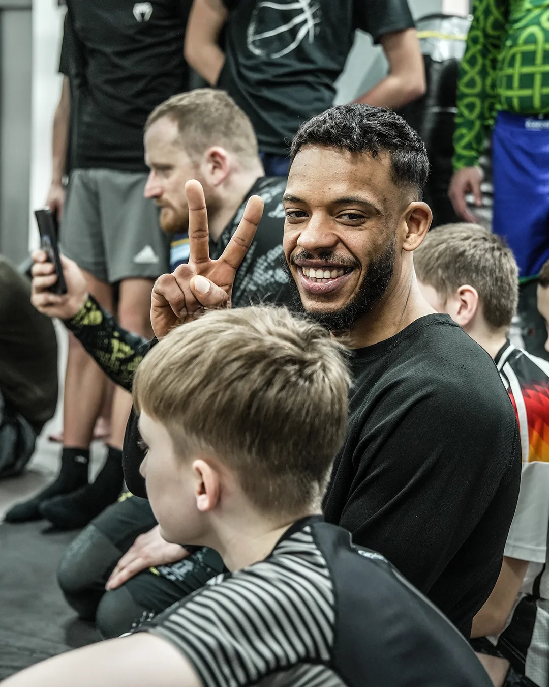 Rund 100 Kinder und Jugendliche auf der Matte: Warum Kampfsport bei GYMRAY in Bamberg so gut ankommt 11 Head Coach Ray lacht und zeigt das Peace-Zeichen, umgeben von Kindern beim Kampfsporttraining bei GYMRAY in Bamberg